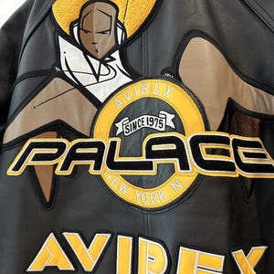 Puede incluir: Chaqueta de cuero negra con parches bordados amarillos y negros. Presenta el texto "PALACE NEW YORK N" y "AVIREX SINCE 1975". Incluye un gr&aacute;fico de dibujos animados en la parte superior de la espalda.