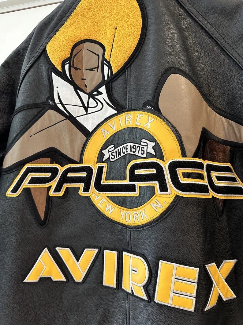 Puede incluir: Chaqueta de cuero negra con parches bordados amarillos y negros. Presenta el texto "PALACE NEW YORK N" y "AVIREX SINCE 1975". Incluye un gr&aacute;fico de dibujos animados en la parte superior de la espalda.