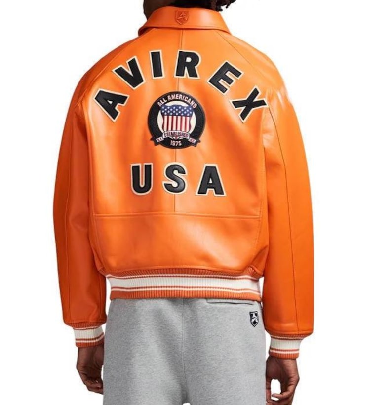 K&ouml;nnte beinhalten: Orangefarbene Lederjacke mit dem Wort "AVIREX" in schwarzen Blockbuchstaben auf dem R&uuml;cken. Darunter ein Aufn&auml;her mit "ALL AMERICANS" und "USA". Die Jacke hat wei&szlig;-rot gestreifte B&uuml;ndchen und Saum. Klassischer Bomber-Stil.