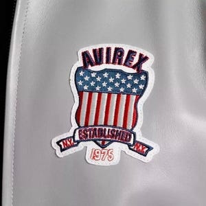 Puede incluir: Primer plano de una chaqueta de cuero gris con un parche cosido. El parche presenta la palabra "AVIREX" en rojo, un dise&ntilde;o de escudo con rayas rojas y blancas, estrellas azules y el texto "ESTABLISHED 1975 NY".