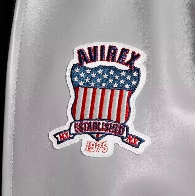 Puede incluir: Primer plano de una chaqueta de cuero gris con un parche cosido. El parche presenta la palabra "AVIREX" en rojo, un dise&ntilde;o de escudo con rayas rojas y blancas, estrellas azules y el texto "ESTABLISHED 1975 NY".