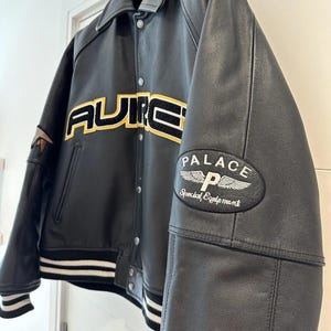 Puede incluir: Chaqueta de cuero negra con la palabra "AURE" en letras amarillas y negras en el pecho. La chaqueta tiene un ribete a rayas blancas y negras en los pu&ntilde;os y el dobladillo. Un parche en la manga dice "PALACE Special Equipment".