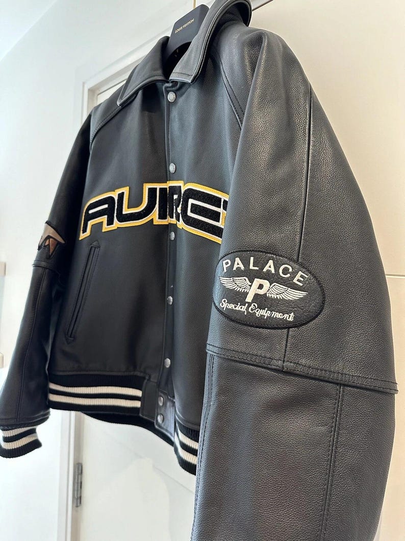 Puede incluir: Chaqueta de cuero negra con la palabra "AURE" en letras amarillas y negras en el pecho. La chaqueta tiene un ribete a rayas blancas y negras en los pu&ntilde;os y el dobladillo. Un parche en la manga dice "PALACE Special Equipment".