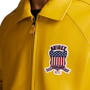 May include: Mustard yellow leather jacket with a collared neckline and a zipper closure. Features a patch with the word "AVIREX" above a design of the American flag and the words "ESTABLISHED 1975" on a banner.