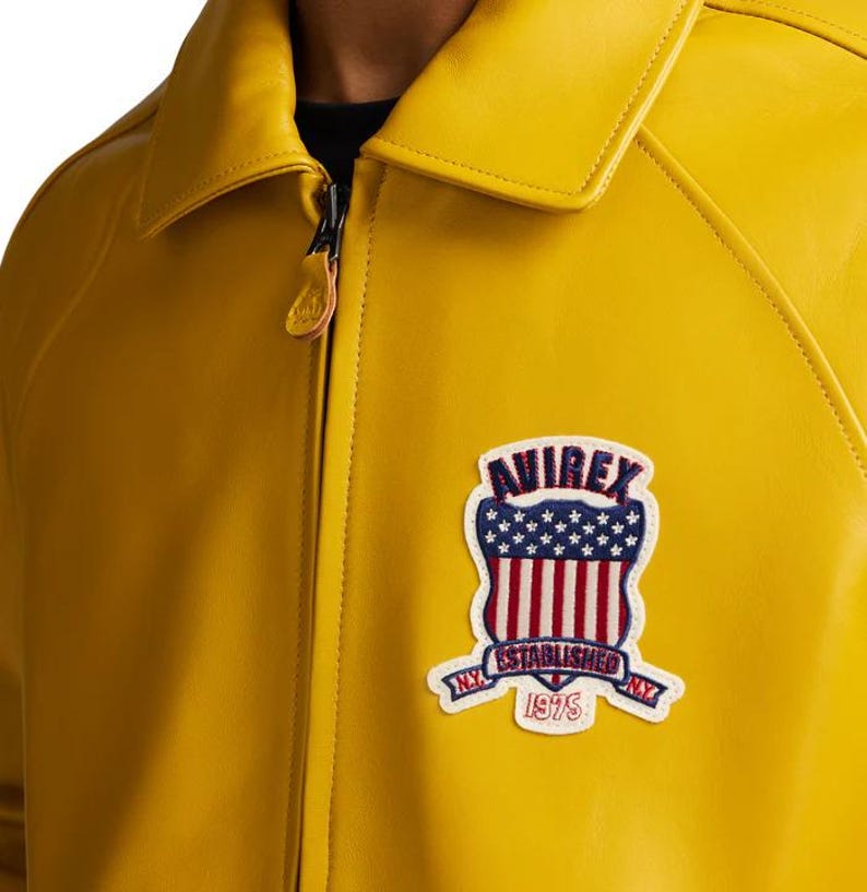 May include: Mustard yellow leather jacket with a collared neckline and a zipper closure. Features a patch with the word "AVIREX" above a design of the American flag and the words "ESTABLISHED 1975" on a banner.