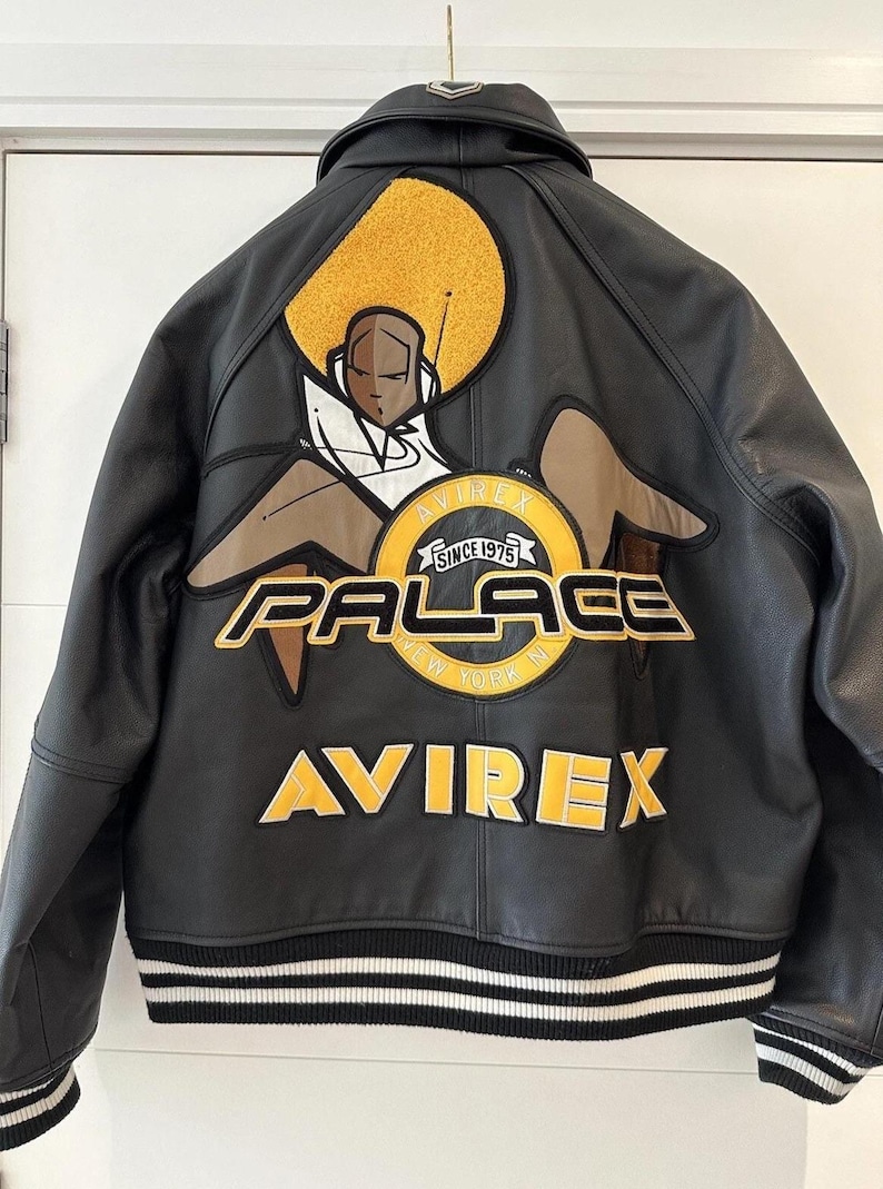 Puede incluir: Chaqueta de cuero negra con un gran gr&aacute;fico en la espalda que presenta una figura sentada, un sol amarillo y las palabras "PALACE NEW YORK IN" y "AVIREX SINCE 1975". La chaqueta tiene un dobladillo a rayas.