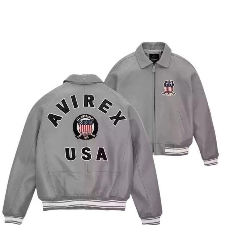 Puede incluir: Chaqueta bomber gris con el texto "AVIREX USA" en la espalda y un parche en la parte delantera. La chaqueta tiene cuello, cierre de cremallera y pu&ntilde;os y dobladillo a rayas blancas y grises. El parche presenta un escudo con rayas y estrellas rojas y blancas.