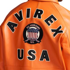 K&ouml;nnte beinhalten: Orangefarbene Lederjacke mit schwarz-wei&szlig;en Buchstaben "AVIREX" und "USA". Ein kreisf&ouml;rmiger Aufn&auml;her zeigt die amerikanische Flagge und die Worte "ALL AMERICANS ESTABLISHED 1975".