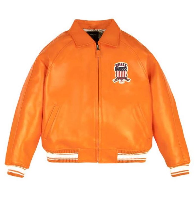 K&ouml;nnte beinhalten: Leuchtend orangefarbene Lederjacke mit Kragen und Rei&szlig;verschluss. Auf der Brust befindet sich ein Aufn&auml;her mit Schilddesign und dem Wort "AVIREX". Die &Auml;rmel und der Saum haben wei&szlig;-orangefarbene Streifen.