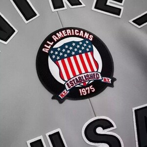 Puede incluir: Primer plano de una chaqueta gris con un parche circular. El parche presenta un dise&ntilde;o de escudo con rayas rojas y blancas, estrellas azules y las palabras "ALL AMERICANS ESTABLISHED N.Y. 1975". Tambi&eacute;n son visibles parches de letras en blanco y negro.