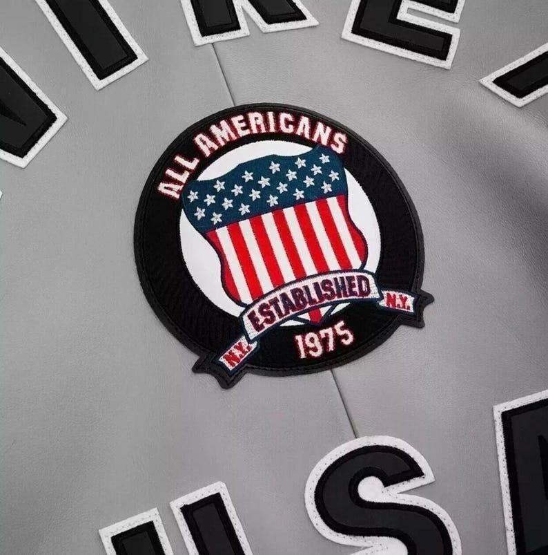 Puede incluir: Primer plano de una chaqueta gris con un parche circular. El parche presenta un dise&ntilde;o de escudo con rayas rojas y blancas, estrellas azules y las palabras "ALL AMERICANS ESTABLISHED N.Y. 1975". Tambi&eacute;n son visibles parches de letras en blanco y negro.