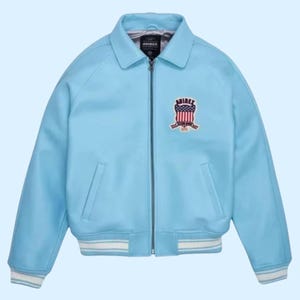 Puede incluir: Chaqueta bomber azul claro con cremallera, cuello y dos bolsillos delanteros. La chaqueta tiene un ribete a rayas blancas y azul claro en los pu&ntilde;os y el dobladillo. Un parche bordado con la palabra "AVIREX" y un dise&ntilde;o patri&oacute;tico est&aacute; en el pecho.