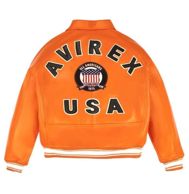 K&ouml;nnte beinhalten: Orangefarbene Lederjacke mit wei&szlig;en und schwarzen Akzenten. Auf der R&uuml;ckseite steht das Wort "AVIREX" &uuml;ber einem runden Aufn&auml;her mit der Aufschrift "ALL AMERICANS ESTABLISHED 1975" und einem US-Flaggen-Design. Darunter steht das Wort "USA".