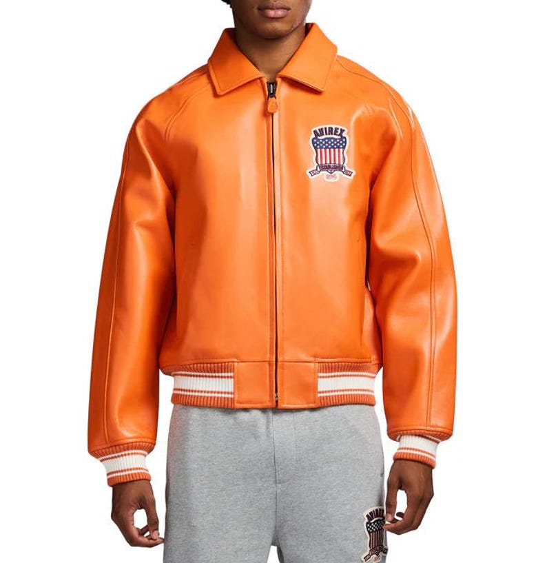 K&ouml;nnte beinhalten: Orangefarbene Lederjacke mit Rei&szlig;verschluss, Kragen und gerippten B&uuml;ndchen und Saum. Sie hat einen Aufn&auml;her auf der Brust mit dem Text "AVIREX" und einem patriotischen Design. Die Jacke hat einen klassischen Bomberstil.