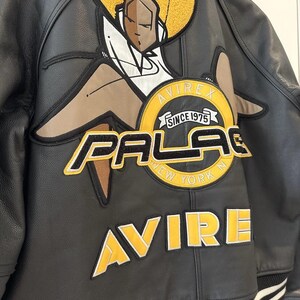 Puede incluir: Chaqueta de cuero negra con un gran dise&ntilde;o bordado en la espalda. El dise&ntilde;o presenta una figura estilizada con un afro amarillo, las palabras "PALACE NEW YORK" y "AVIREX SINCE 1975" en amarillo y negro.