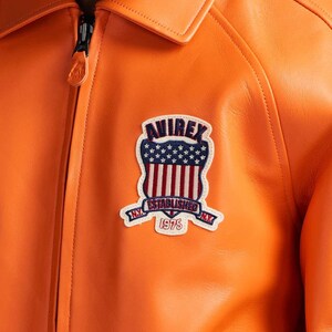 K&ouml;nnte beinhalten: Nahaufnahme einer orangefarbenen Lederjacke mit schwarzem Rei&szlig;verschluss und Kragen. Die Jacke hat einen Aufn&auml;her mit dem Text "AVIREX ESTABLISHED 1975" und einem Design der amerikanischen Flagge.