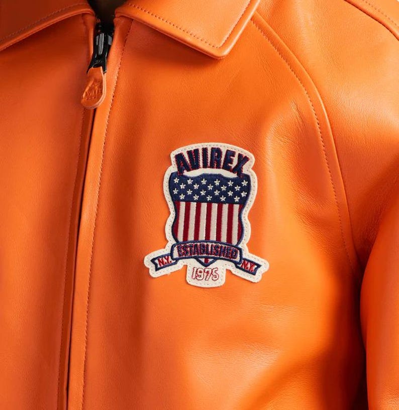 K&ouml;nnte beinhalten: Nahaufnahme einer orangefarbenen Lederjacke mit schwarzem Rei&szlig;verschluss und Kragen. Die Jacke hat einen Aufn&auml;her mit dem Text "AVIREX ESTABLISHED 1975" und einem Design der amerikanischen Flagge.