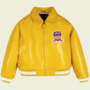 May include: A bright yellow leather jacket with a bomber style. The jacket features a white and yellow striped trim on the cuffs and hem. An embroidered patch with a shield design and the word "AVIREX" is on the chest.