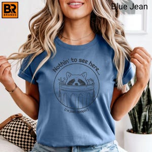 May include: Blue jean t-shirt with a raccoon graphic inside a trash can, with the text "Nothin' to see here..." and "(I'm just goopin')". The shirt has short sleeves and a crew neck.
