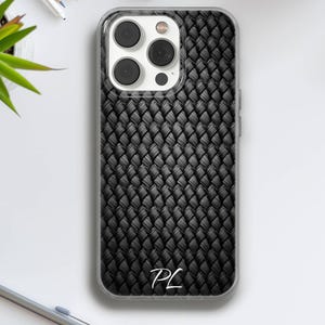 Op de afbeelding: Zwarte telefoonhoes met een geweven textuurontwerp. De hoes heeft een doorzichtige rand en toont de initialen "PL" in het wit aan de onderkant. De telefoonhoes ligt op een wit oppervlak met kantoorbenodigdheden op de achtergrond.