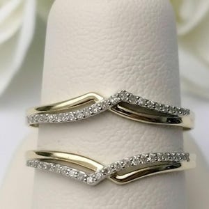 May include: Two gold-toned, curved rings with a row of small, sparkling diamonds set along the top curve. The rings are displayed on a white surface, with a blurred white flower in the background. The rings are likely wedding bands or engagement rings.
