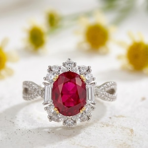 May include: A silver-toned ring featuring a large oval-cut deep red gemstone at the centre, surrounded by a halo of small clear baguette and round-cut stones. The band has a twisted design encrusted with tiny clear stones on both sides.