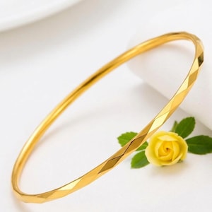 May include: A gold-colored bangle bracelet with a geometric, faceted design. The bracelet is circular and sits on a white surface, next to a yellow rose with green leaves. The bangle is a simple, elegant piece of jewelry.