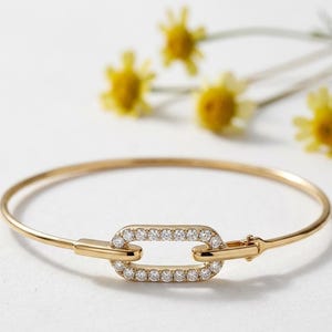 May include: A gold-colored bangle bracelet with a rectangular, diamond-studded clasp. The bracelet is set against a white background with yellow flowers in the background. The bracelet is a delicate piece of jewelry.