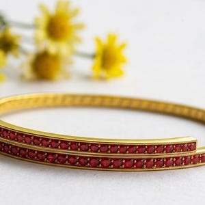 May include: Gold-toned bangle bracelet with two rows of small, red gemstones. The bracelet is set against a white background with yellow flowers in the background. A piece of jewelry.