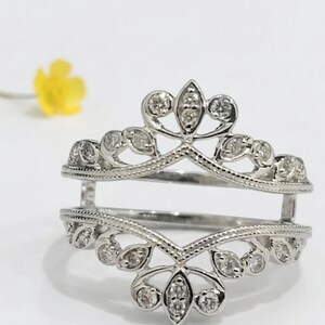 Op de afbeelding: Een zilveren ringbeschermer met een bloemen- en bladerontwerp, bezet met kleine, heldere edelstenen. De ring heeft een open ontwerp, met twee banden verbonden door verticale steunen. Een gele bloem is wazig op de achtergrond.