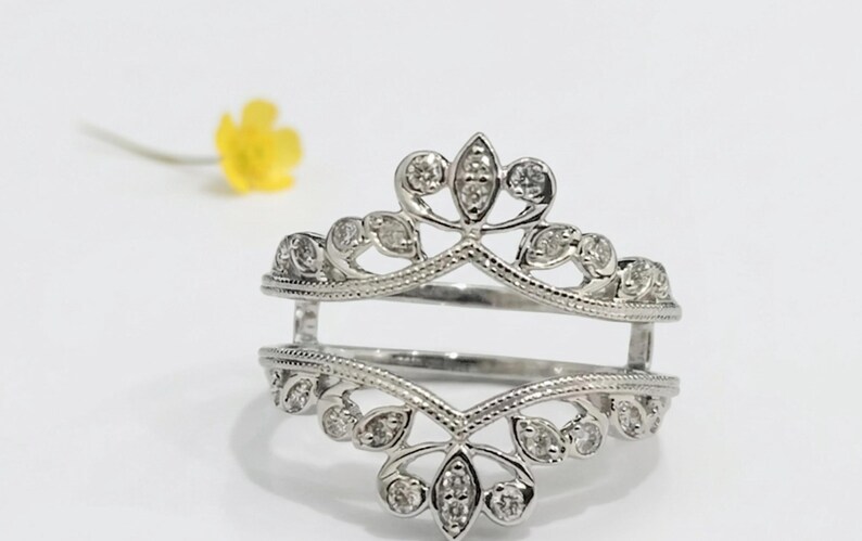 Op de afbeelding: Een zilveren ringbeschermer met een bloemen- en bladerontwerp, bezet met kleine, heldere edelstenen. De ring heeft een open ontwerp, met twee banden verbonden door verticale steunen. Een gele bloem is wazig op de achtergrond.