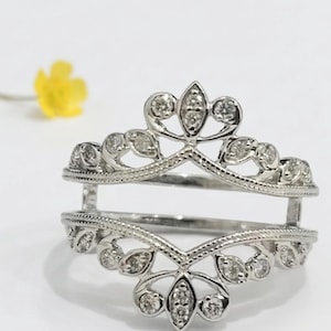 Op de afbeelding: Een zilveren ringbeschermer met een bloemen- en bladerontwerp, bezet met kleine, heldere edelstenen. De ring heeft een open ontwerp, met twee parallelle banden verbonden door verticale steunen. Een gele bloem is wazig op de achtergrond.