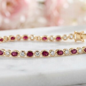 May include: A gold tennis bracelet featuring alternating oval-cut red rubies and round-cut clear diamonds in bezel settings. The bracelet is constructed with a series of linked metal segments and includes a secure box clasp closure mechanism.