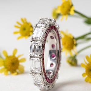 May include: A diamond eternity ring featuring emerald-cut diamonds and red gemstones. The ring, set in white metal, is shown close-up, with yellow flowers in the background.