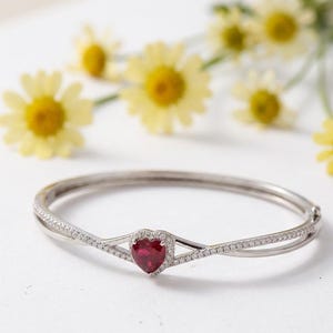 May include: A silver bracelet with a heart-shaped, ruby-colored center stone. The bracelet features a delicate design with small, clear stones and is set against a backdrop of yellow flowers.