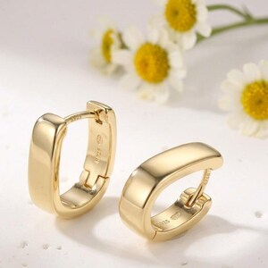 May include: A pair of gold-colored, rectangular hoop earrings. The earrings have a smooth, polished surface and a hinged closure. The background features white flowers with yellow centers.
