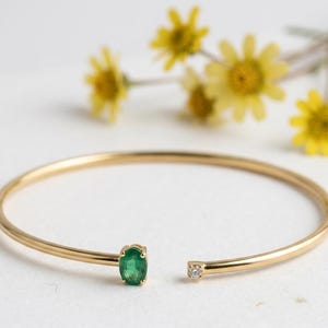 Op de afbeelding: Een open armband in goudkleur met een ovale smaragdgroene edelsteen en een kleine diamant. De armband staat tegen een witte achtergrond met gele bloemen.