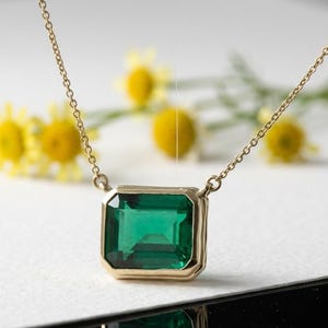 Può includere: Una collana d'oro con un pendente di smeraldo verde intenso di forma rettangolare. Il pendente è incastonato in una cornice d'oro e sospeso a una sottile catena d'oro. Fiori gialli sullo sfondo aggiungono un tocco di colore.