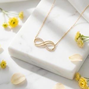 May include: A gold-toned necklace featuring an infinity symbol pendant. The pendant is accented with small, clear, sparkling stones along one side of the loop. The delicate chain is attached to both ends of the infinity-shaped metal centerpiece.
