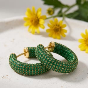 May include: A pair of gold hoop earrings featuring a thick, rounded design fully encrusted with small, vibrant green stones. The earrings have a polished gold metal post and hinge mechanism, showcasing a dense, textured surface of green gems.