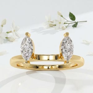 Puede incluir: Un anillo de oro con dos diamantes talla marquesa. Los diamantes están engastados a ambos lados de la banda del anillo, creando un diseño único. El anillo se muestra sobre una superficie blanca, con flores blancas en el fondo.