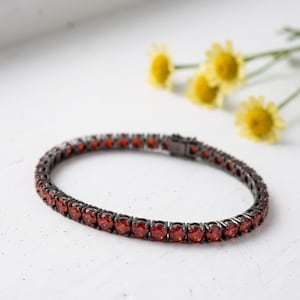 May include: A tennis bracelet featuring a series of round, faceted, deep red stones set in a dark, gunmetal-toned metal frame. The bracelet is designed with a secure box clasp mechanism and displays a continuous row of vibrant red gemstones.