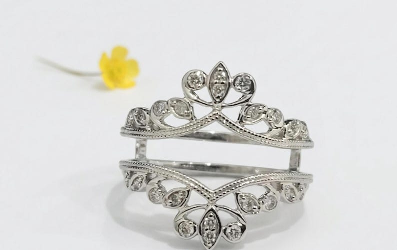 Op de afbeelding: Een zilveren ringbeschermer met een bloemen- en bladerontwerp, bezet met kleine, heldere edelstenen. De ring heeft een open ontwerp, met twee banden verbonden door verticale steunen. Een gele bloem is wazig op de achtergrond.