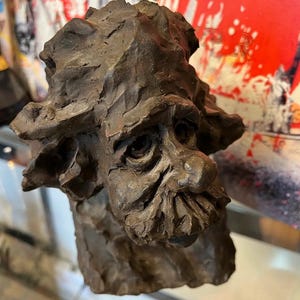 May include: A brown ceramic sculpture of a person's head with a textured surface. The sculpture has a detailed face with a long nose and a beard.