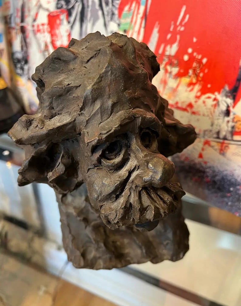 May include: A brown ceramic sculpture of a person's head with a textured surface. The sculpture has a detailed face with a long nose and a beard.