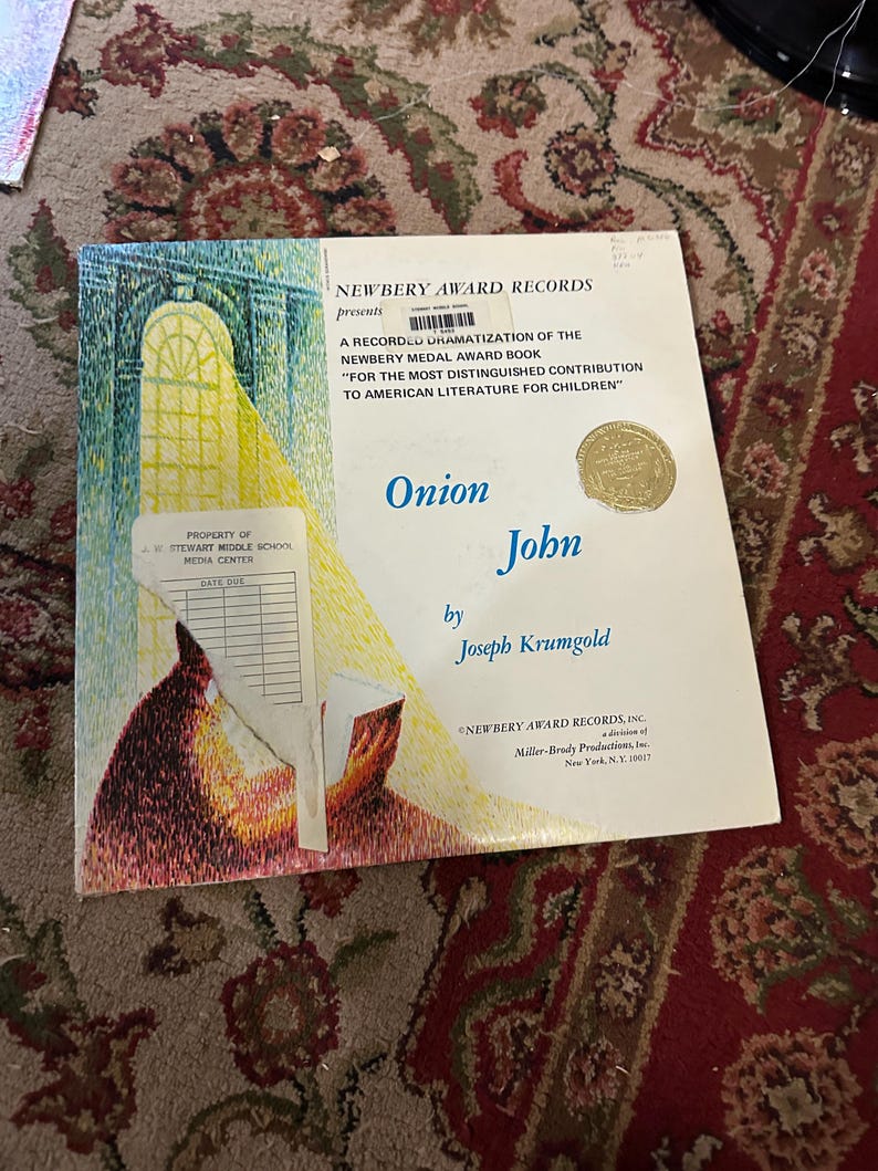 May include: A record dramatization of the Newbery Medal Award book "Onion John" by Joseph Krumgold.  The cover features a colorful illustration of a boy reading a book.  The book is presented by Newbery Award Records, Inc.
