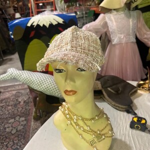 May include: A beige tweed newsboy cap with a frayed brim sits on a mannequin head. The mannequin wears a layered pearl necklace. Other items in the background include a pink dress, a decorative chair, and a gold-colored object.