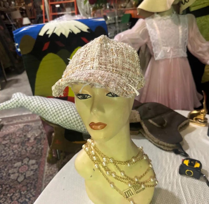 May include: A beige tweed newsboy cap with a frayed brim sits on a mannequin head. The mannequin wears a layered pearl necklace. Other items in the background include a pink dress, a decorative chair, and a gold-colored object.