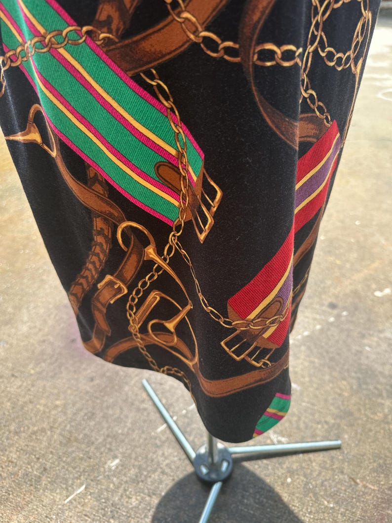 May include: Black fabric with a repeating pattern of gold chains, brown leather straps, and colorful striped ribbons. The fabric has a textured surface.