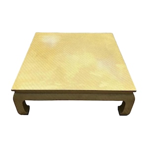 May include: A square, low-profile table with a light yellow, textured surface and a subtle, repeating pattern. The table features four curved legs that extend outward from the corners, providing a stable base for the flat, rectangular tabletop.